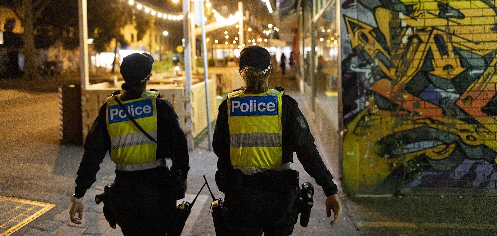 Two female police officers are walking down the street at night. They are facing away from us, and you can clearly read their high vis vests marked 'Police'. The street is brightly lit and you can see some graffiti on the wall next to them. 