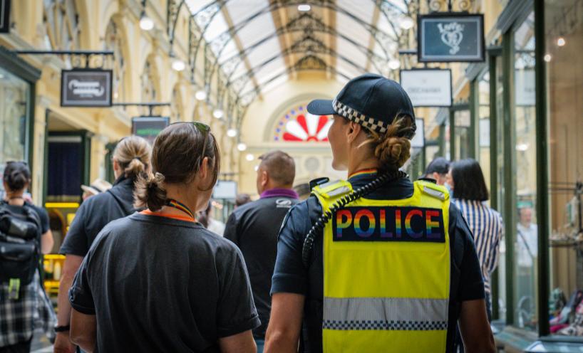 From the back - officer with Victoria Police pride logo high vis vest on and a member of public walking inside a shopping centre.  