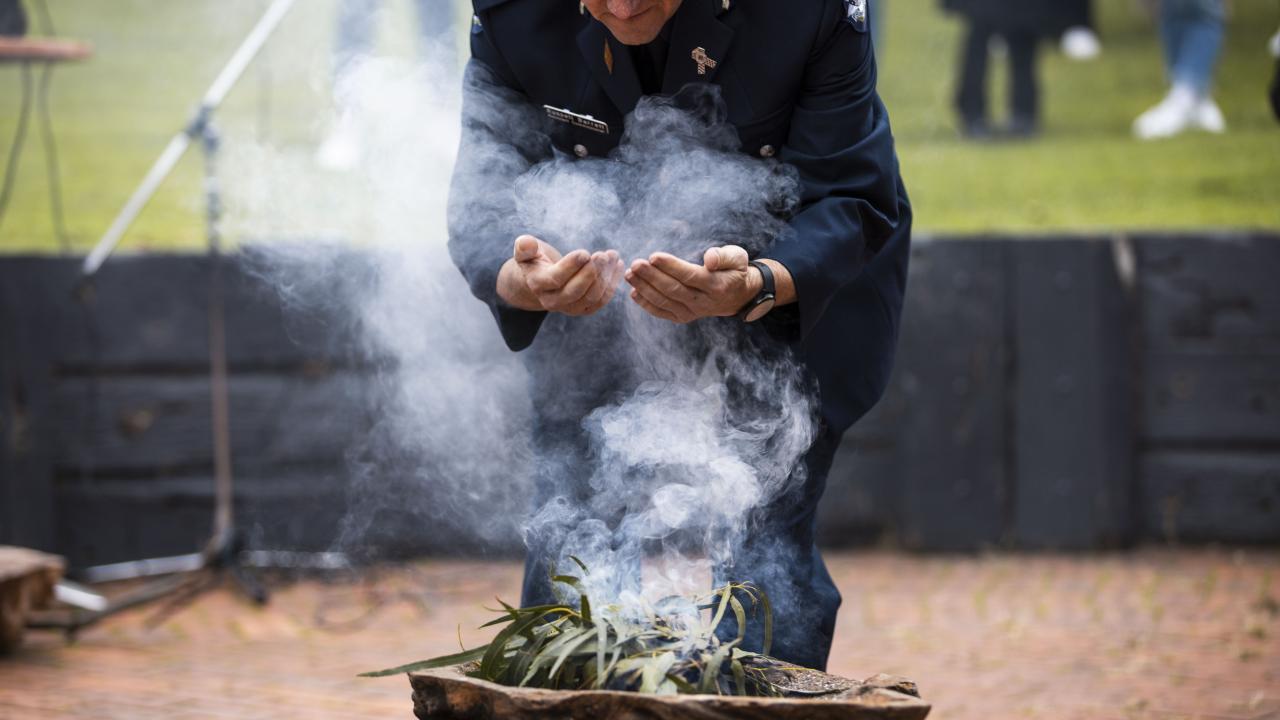 A uniformed police officer participating in an outdoor smoking ceremony, scooping the smoke with his hands, with event attendees in the background.