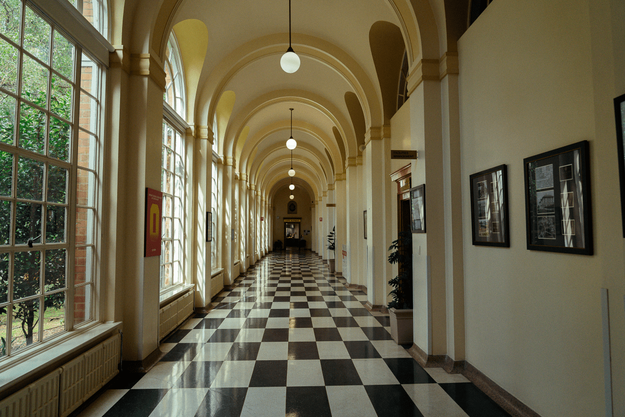 Inside the halls of the Victoria Police Academy. There is a high arched ceiling with an art deco feel,  black and white tiled floors, pictures down one wall and on the other side, large pane glass windows looking out into the garden.