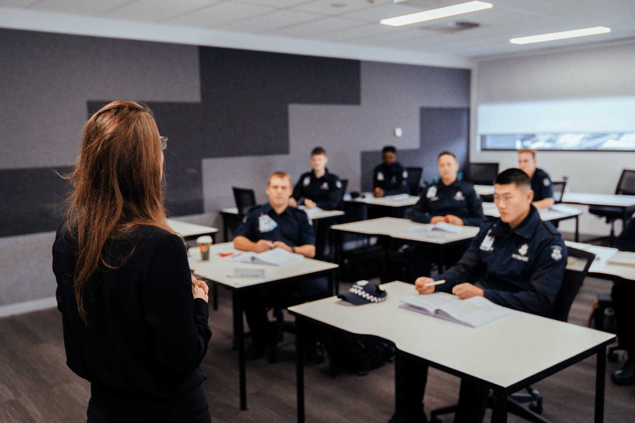 Victoria Police recruits take a class at the Academy. There is an instructor standing at the front of the classroom, speaking to them as they take notes at their desks. 