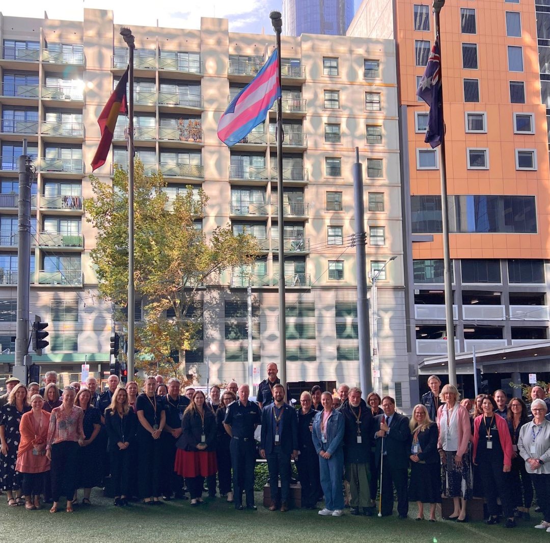 Members of the community stand under the raised Trans flag, Aboriginal flag, and Australian flag. 