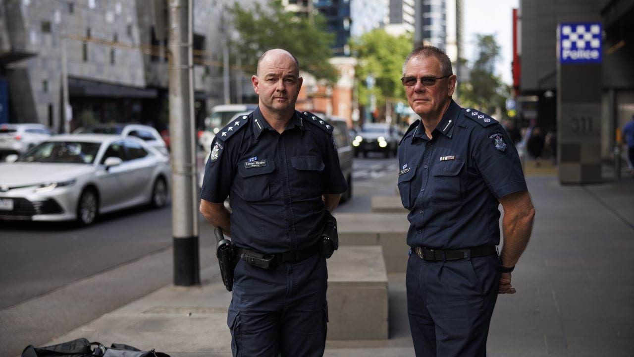 Two officers in police uniform stand in front of the Victoria Police Centre. 